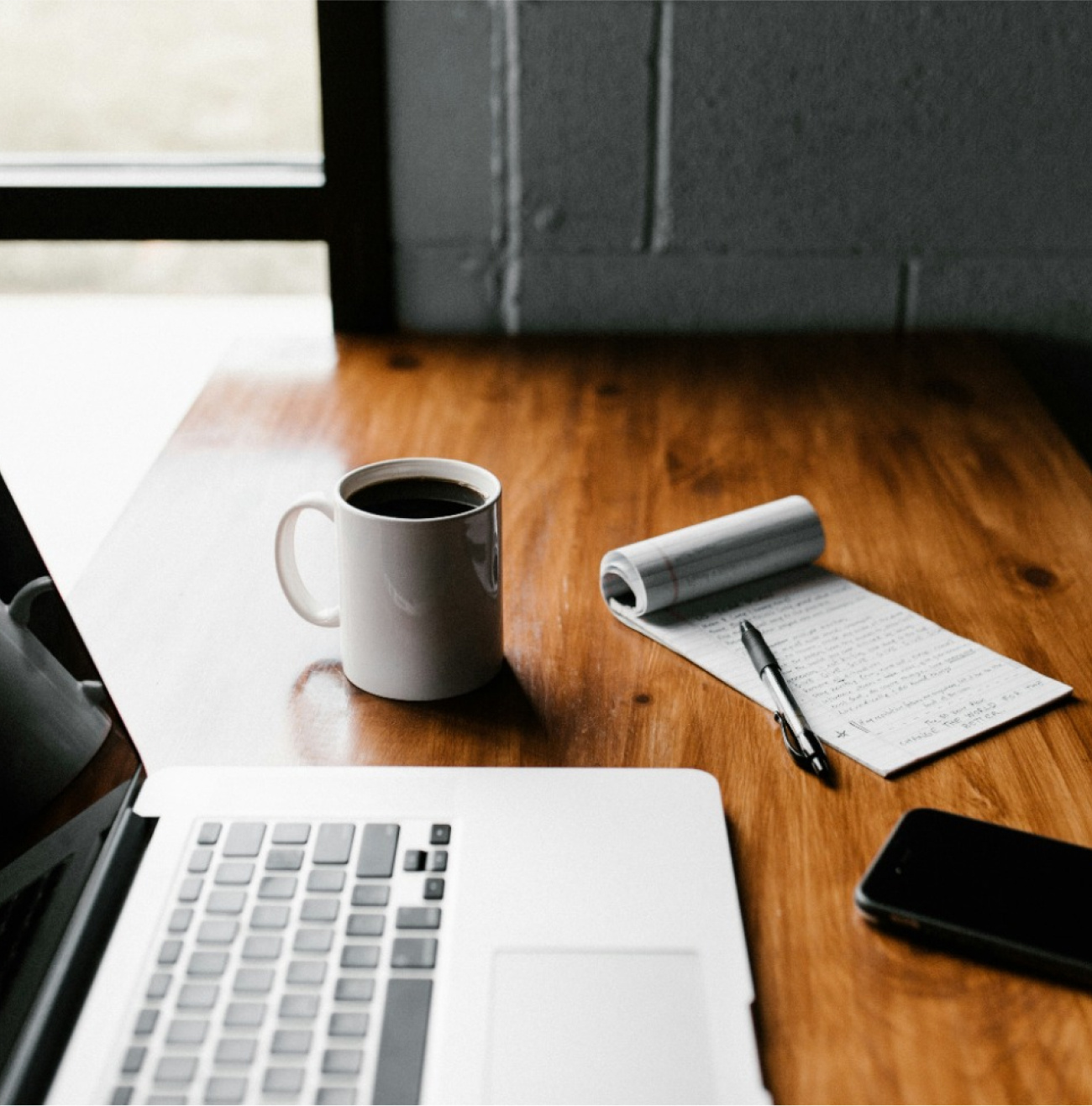 A warm workspace with coffee, a laptop, handwritten notes, and a phone, set on a wooden table by the window